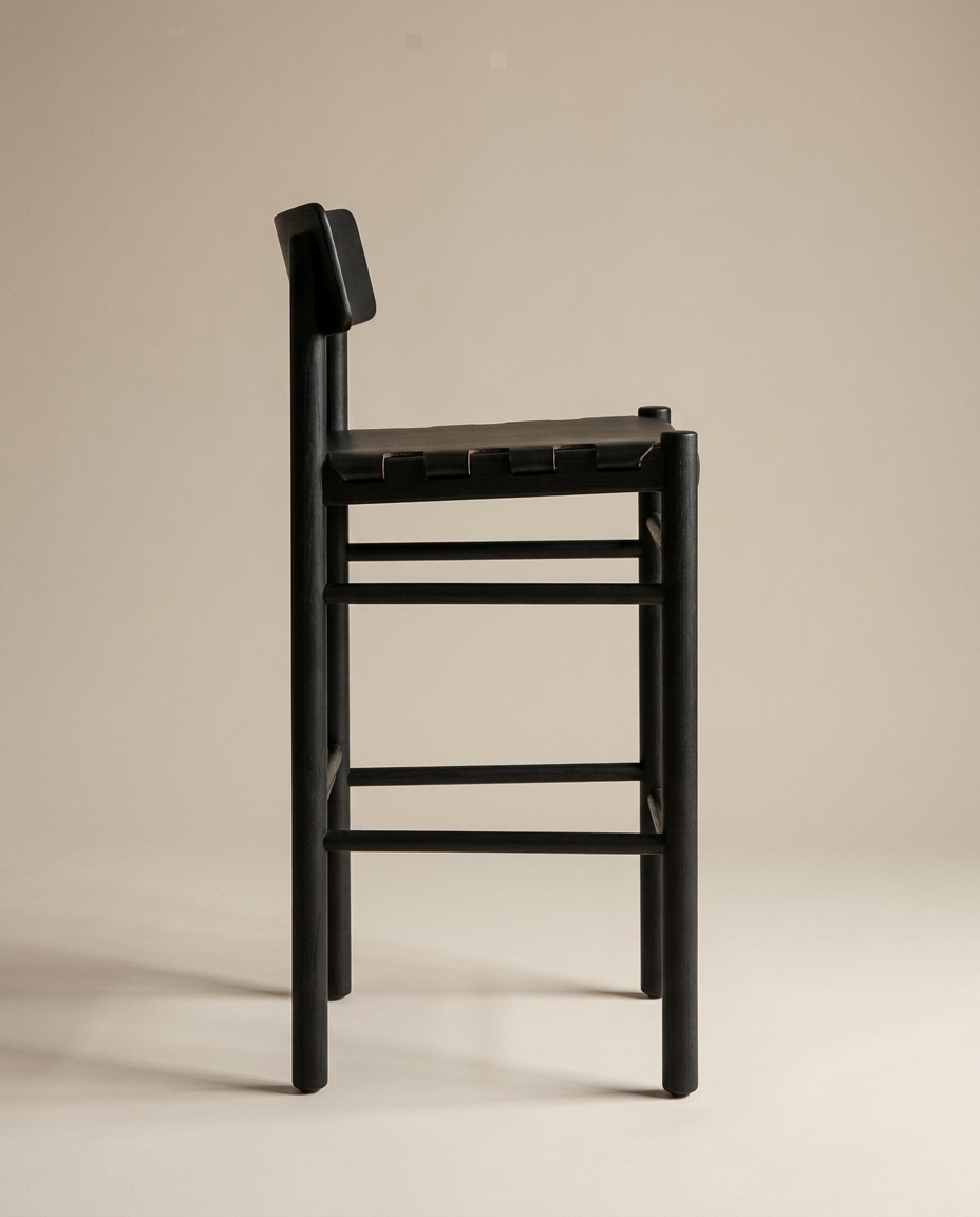 The Cesco Counter Stool - Black Ash by SOBU is crafted from solid American ash and showcases a black wooden frame with a tall backrest and minimalist, modern aesthetic. Photographed from the side against a plain beige background, this stool features straight lines, no armrests, and an elegantly finished top grain leather seat.