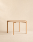 The Cove Dining Table | Light Oak by Wooden Stories features a simple, round design expertly crafted from solid American oak, supported by four sturdy legs. Showcased against a neutral beige background, its contract-grade construction offers lasting style and durability.
