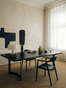 A minimalist office features a black desk and chair, modern lamp, books, and curated decor. Sunlight passes through sheer curtains onto abstract art adorning the beige wall. The Mars Live Edge Dining Table by SOBU—crafted from solid black ash with elegant curved legs—serves as a sophisticated workspace, elevating the room’s calm and refined ambiance.