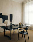 A minimalist office features a black desk and chair, modern lamp, books, and curated decor. Sunlight passes through sheer curtains onto abstract art adorning the beige wall. The Mars Live Edge Dining Table by SOBU—crafted from solid black ash with elegant curved legs—serves as a sophisticated workspace, elevating the room’s calm and refined ambiance.