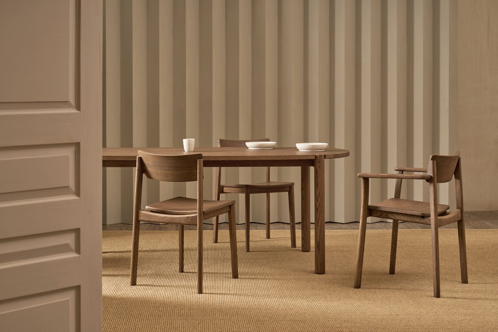 A minimalist dining room with a Tolv wooden table, three Wooden Stories Poise Dining Chairs in Espresso Oak, white bowls and cups on the table, beige carpeting and vertical beige paneling. A partially open white door appears in the foreground.