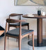 Two SOBU Copenhagen Dining Chairs - Walnut with black leather seats sit by a round wooden table arranged with white plates, a white mug, and a small potted plant. Abstract wall art is visible in the background.