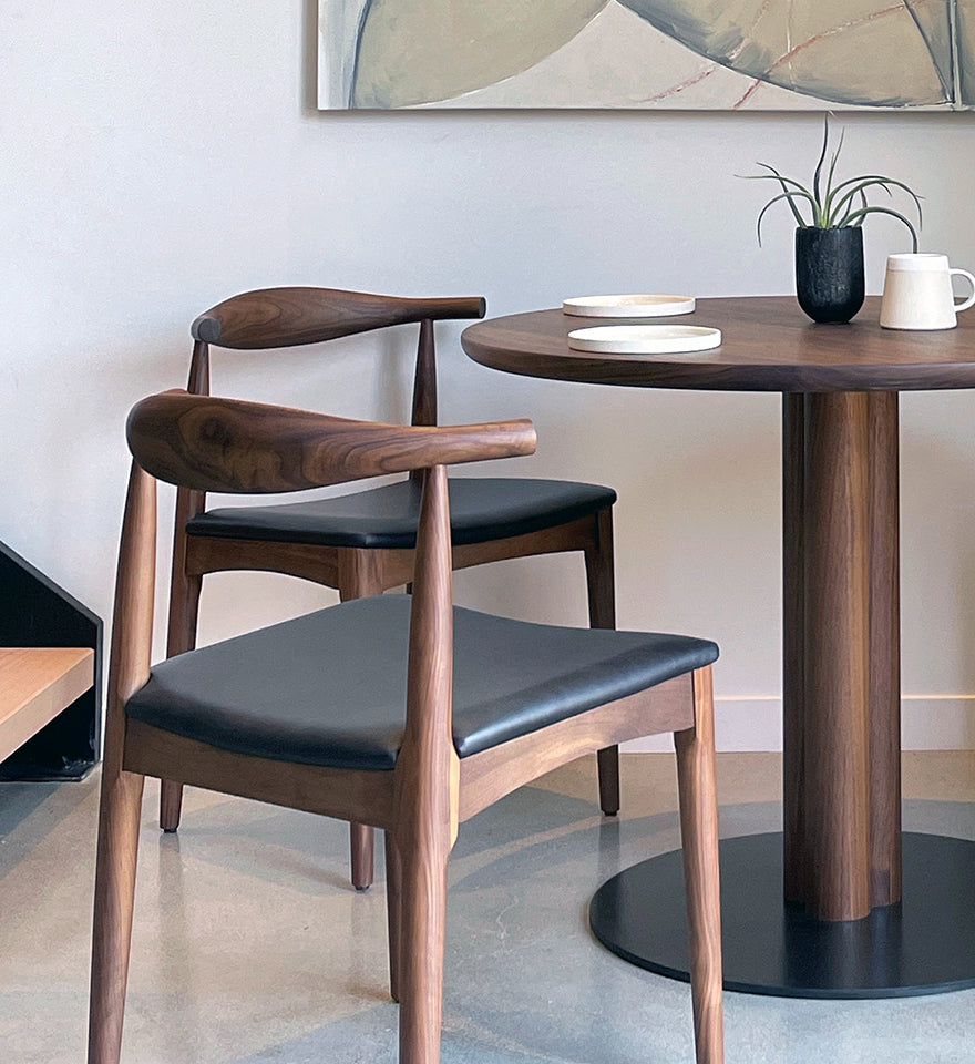 Two SOBU Copenhagen Dining Chairs - Walnut with black leather seats sit by a round wooden table arranged with white plates, a white mug, and a small potted plant. Abstract wall art is visible in the background.