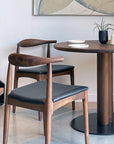 Two SOBU Copenhagen Dining Chairs - Walnut with black leather seats sit by a round wooden table arranged with white plates, a white mug, and a small potted plant. Abstract wall art is visible in the background.