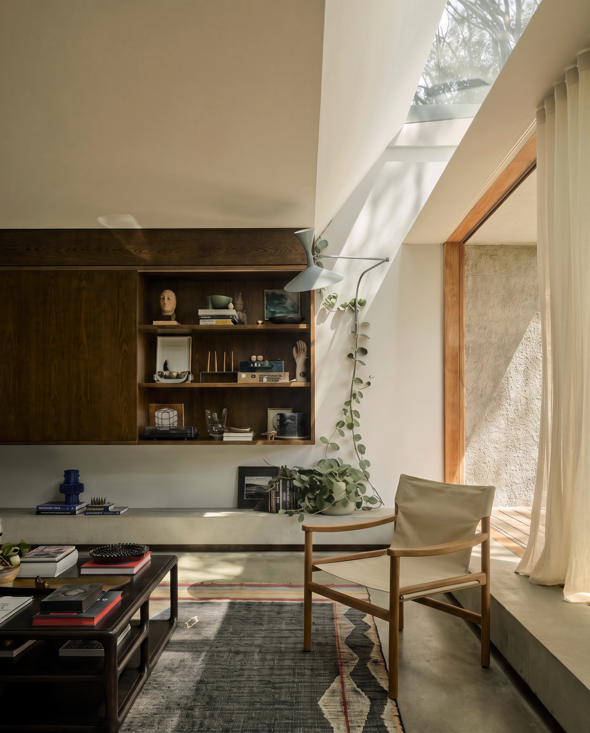 A sunlit modern living room featuring the Ethnicraft Eda Coffee Table, wooden shelves adorned with books and decor pieces, a trailing potted plant, a light wooden chair with European craftsmanship, glass doors opening outside, and sunlight streaming through a skylight.