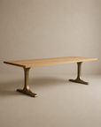 The SOBU Tommaso Dining Table features a rectangular solid wood top and two wide, curved brass T-leg pedestals, set against a plain beige background.
