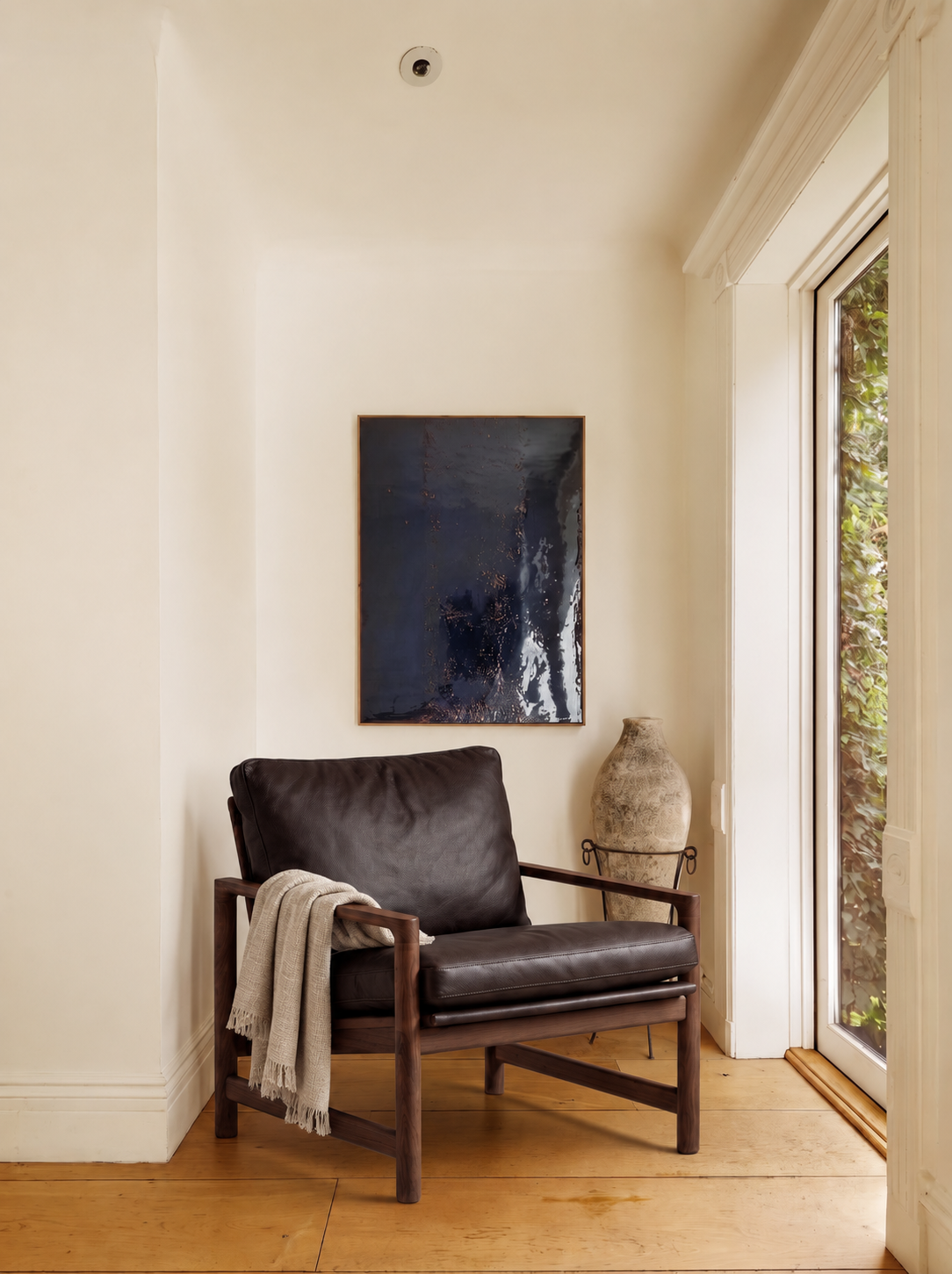 A black Troubadour Lounge Chair | Leather by Wooden Stories with a beige throw sits by a window, dark abstract art hangs behind, and a rustic vase stands nearby in a light-walled, wood-floored room.