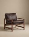 The Troubadour Lounge Chair | Leather by Wooden Stories features a dark brown leather cushion and wooden frame, inspired by Danish design, set against a neutral beige background.