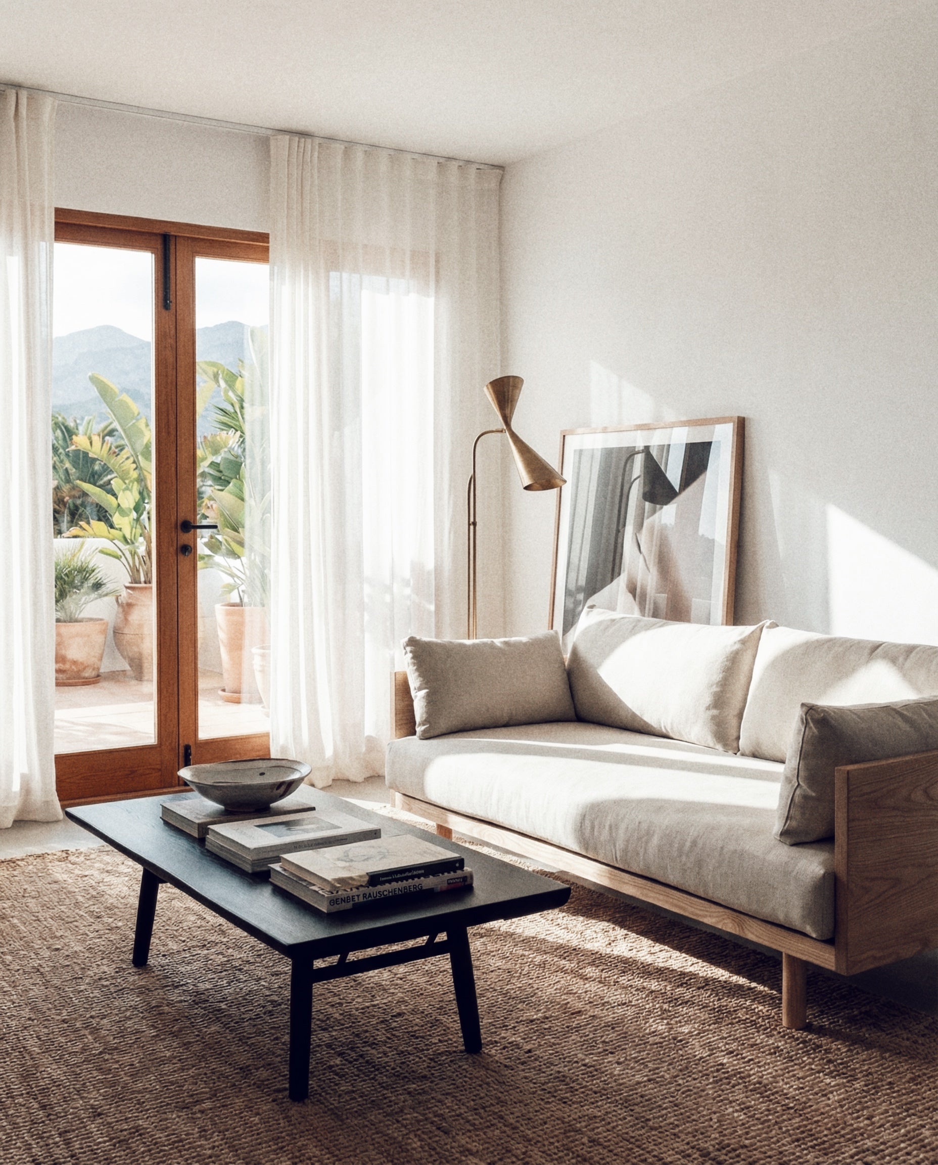 A bright, minimalist living room features the SOBU Woody Sofa with white ash and natural kapok cushions, a black coffee table, woven rug, large framed art, and floor-to-ceiling windows opening to a patio with greenery and mountain views.