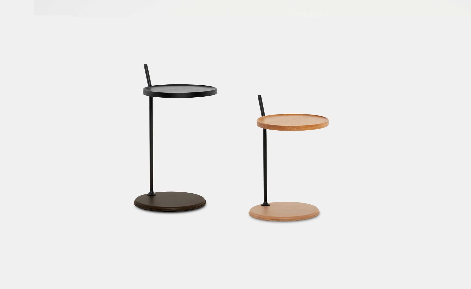 Two Bystander Side Tables by Wooden Stories—both small and round with single central legs and flat circular bases—are shown against a plain white background. One table displays a classic Danish-inspired design in black, while the other features solid wood in a light finish; each is distinguished by its vertical handle.