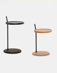 Two Bystander Side Tables by Wooden Stories—both small and round with single central legs and flat circular bases—are shown against a plain white background. One table displays a classic Danish-inspired design in black, while the other features solid wood in a light finish; each is distinguished by its vertical handle.