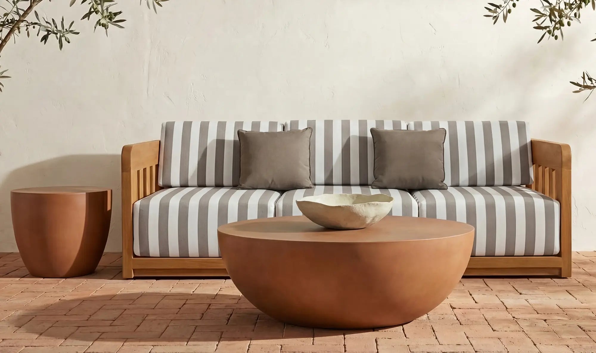 Outdoor patio with a wooden sofa featuring gray and white striped cushions, three gray pillows, the Moes Insitu Coffee Table, a small side table, and a textured bowl on brick flooring against a beige wall—perfect for those who appreciate contemporary home decor.