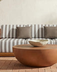 Outdoor patio with a wooden sofa featuring gray and white striped cushions, three gray pillows, the Moes Insitu Coffee Table, a small side table, and a textured bowl on brick flooring against a beige wall—perfect for those who appreciate contemporary home decor.