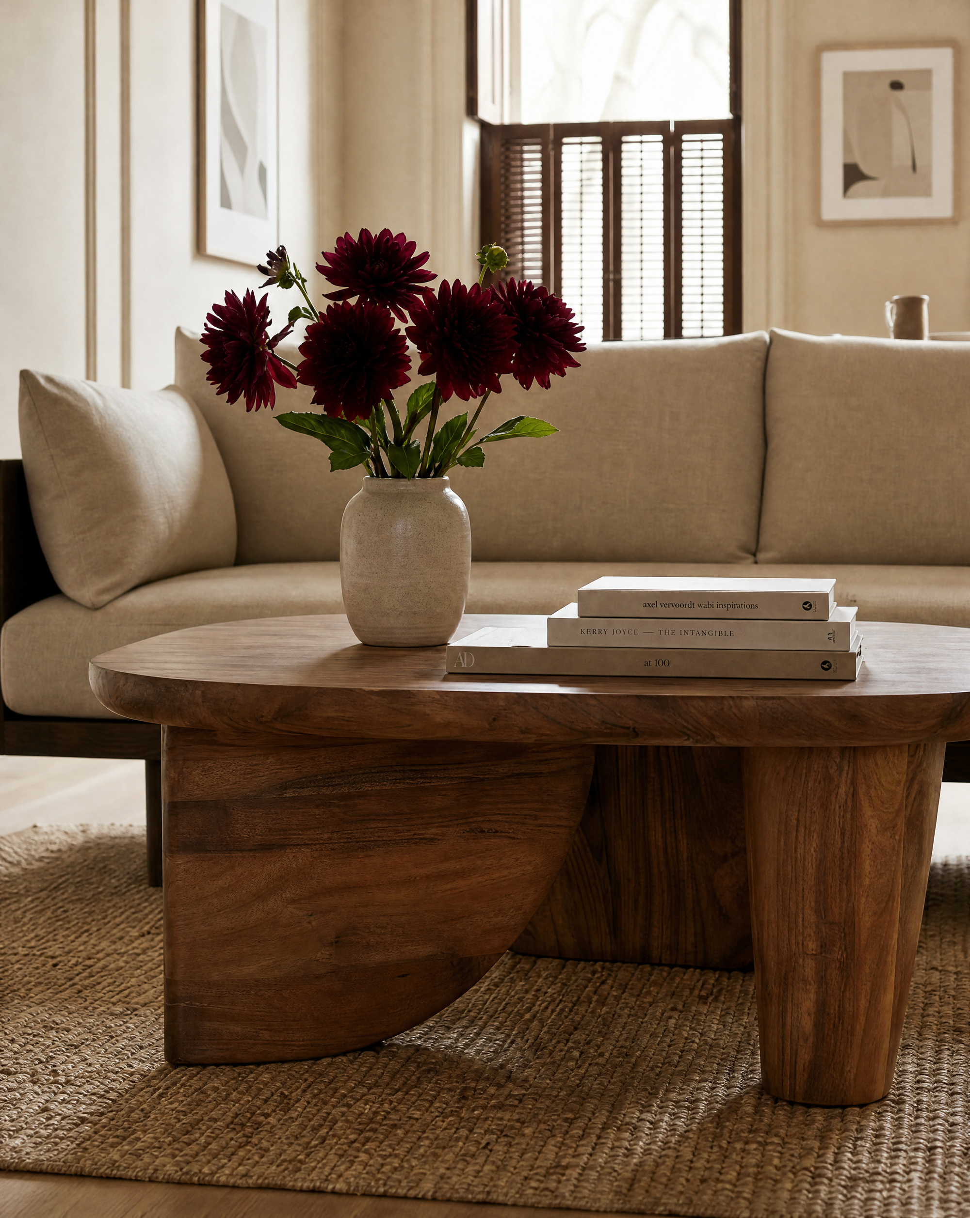 A SOBU Nick Coffee Table, expertly crafted from solid Acacia wood and styled with a vase of dark red flowers alongside a stack of books, rests atop a woven rug before a beige sofa in a softly lit, modern living room—an ideal showcase of modern art furniture that seamlessly merges style and function.