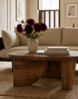 A SOBU Nick Coffee Table, expertly crafted from solid Acacia wood and styled with a vase of dark red flowers alongside a stack of books, rests atop a woven rug before a beige sofa in a softly lit, modern living room—an ideal showcase of modern art furniture that seamlessly merges style and function.