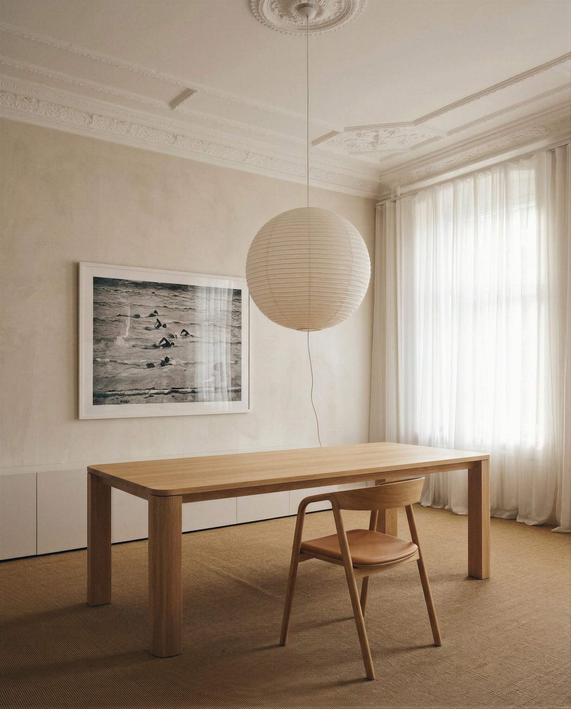 Minimalist dining room featuring the Oli Dining Table by Wooden Stories crafted from solid American oak, paired with a single matching chair. The scene is complemented by a large round paper lantern light, sheer curtains, and an ornate ceiling, with a black-and-white framed photo adorning the wall. These contract-grade furnishings rest within a neutral color palette and benefit from soft lighting for an inviting ambience.