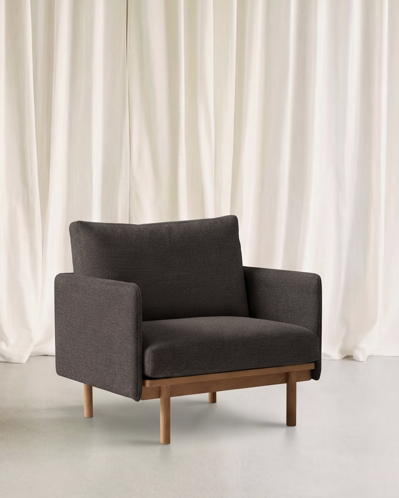 The Pensive Lounge Chair | Fabric by Wooden Stories, in modern dark gray with wooden legs, sits on a light floor before white curtains, showcasing mid-century Danish design elegance.
