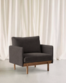 The Pensive Lounge Chair | Fabric by Wooden Stories, in modern dark gray with wooden legs, sits on a light floor before white curtains, showcasing mid-century Danish design elegance.