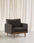The Pensive Lounge Chair | Fabric by Wooden Stories, in modern dark gray with wooden legs, sits on a light floor before white curtains, showcasing mid-century Danish design elegance.