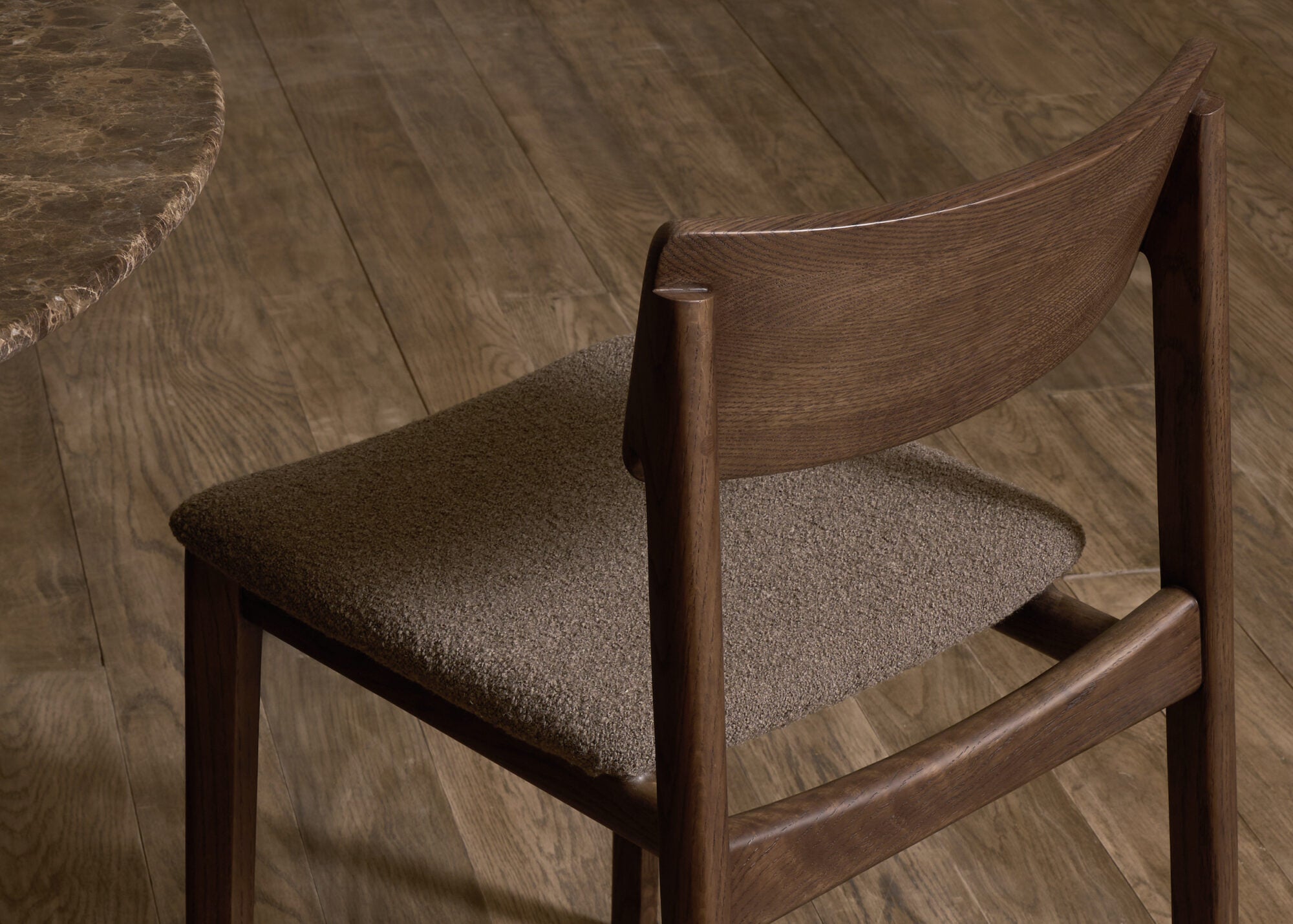 A close-up of the Poise Dining Chair | Espresso Oak with Fabric by Wooden Stories shows its brown, textured seat on a wooden floor beside a round, dark stone table, set within a warm and earthy color palette.