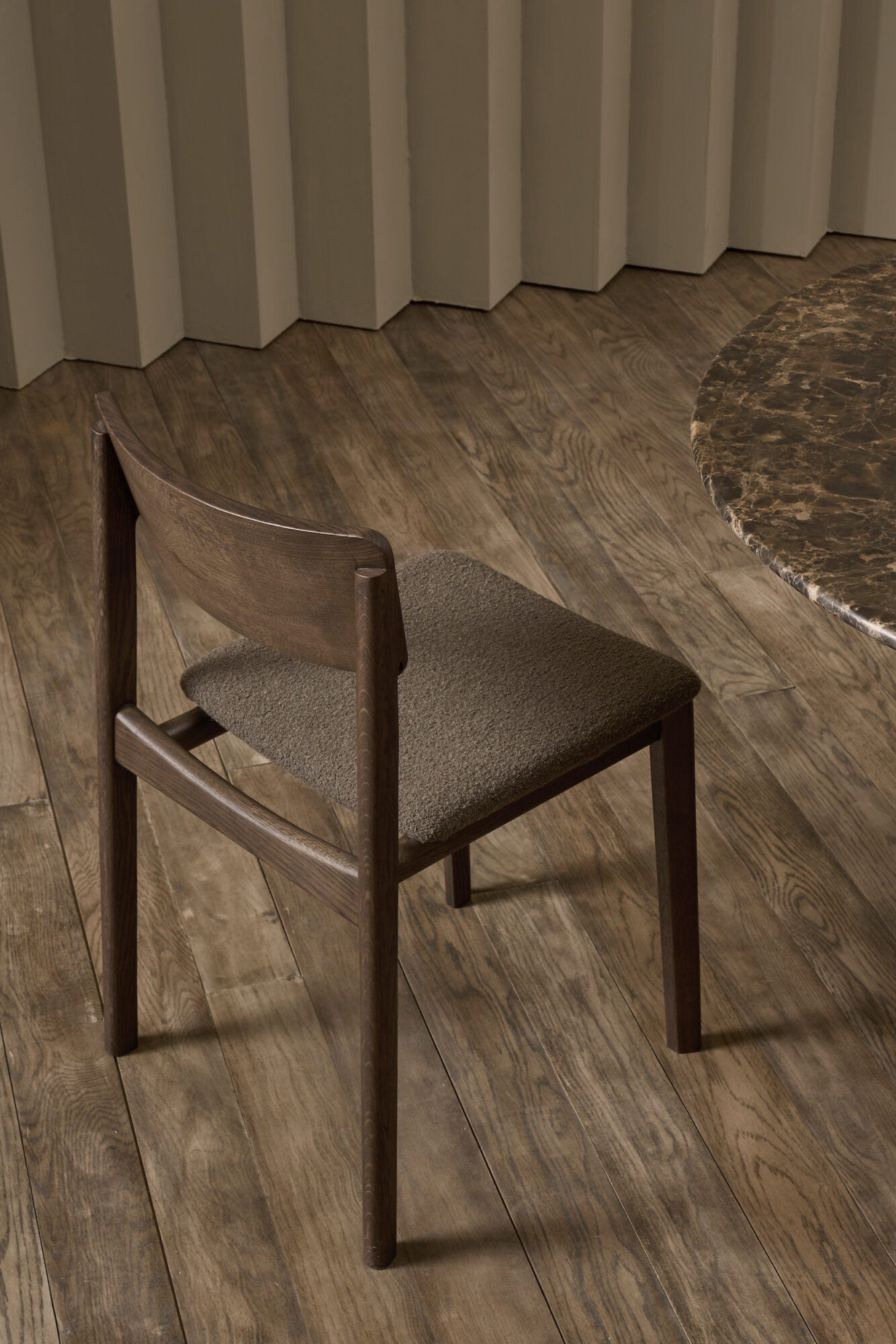 The Poise Dining Chair in Espresso Oak with Fabric by Wooden Stories, designed by Patryk Koca, sits on a wooden floor next to a round marble-topped table, backed by a beige wall with vertical grooves.