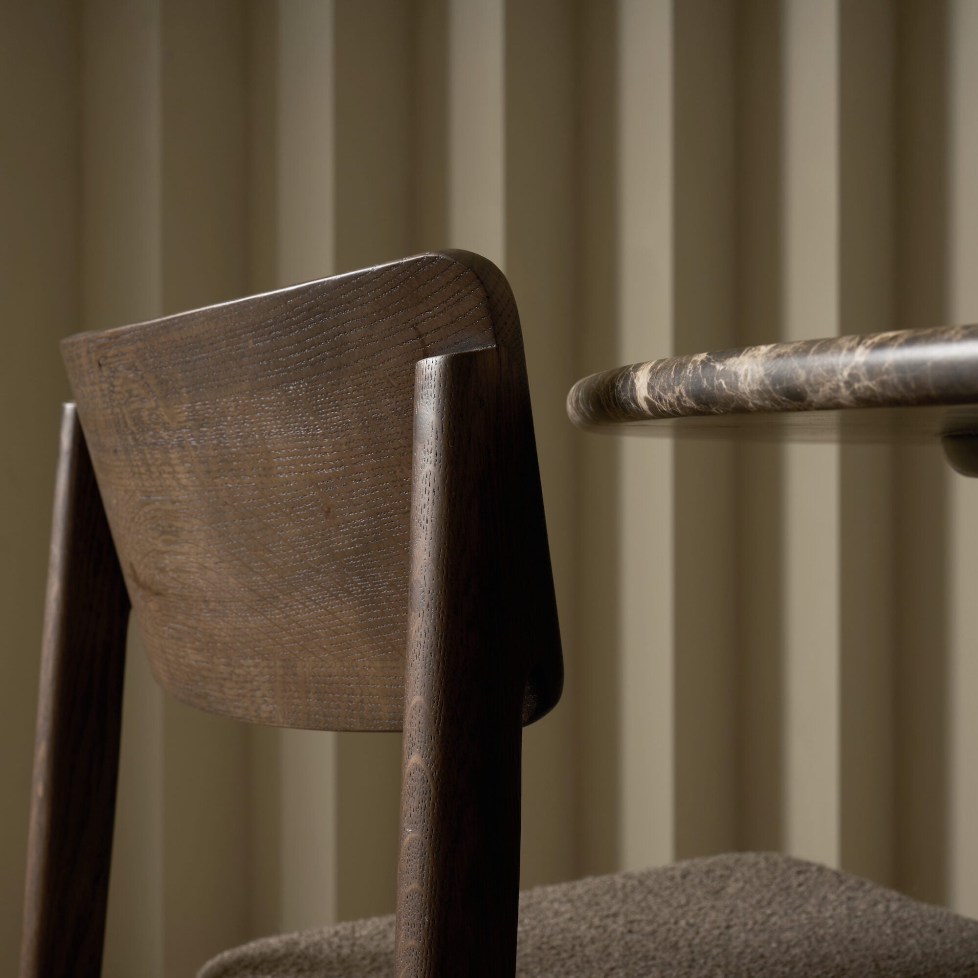 A close-up of the Poise Dining Chair in Espresso Oak with Fabric by Wooden Stories sits beside a marble-edged round table, set against a beige wall with vertical ridges, highlighting textures and refined materials.