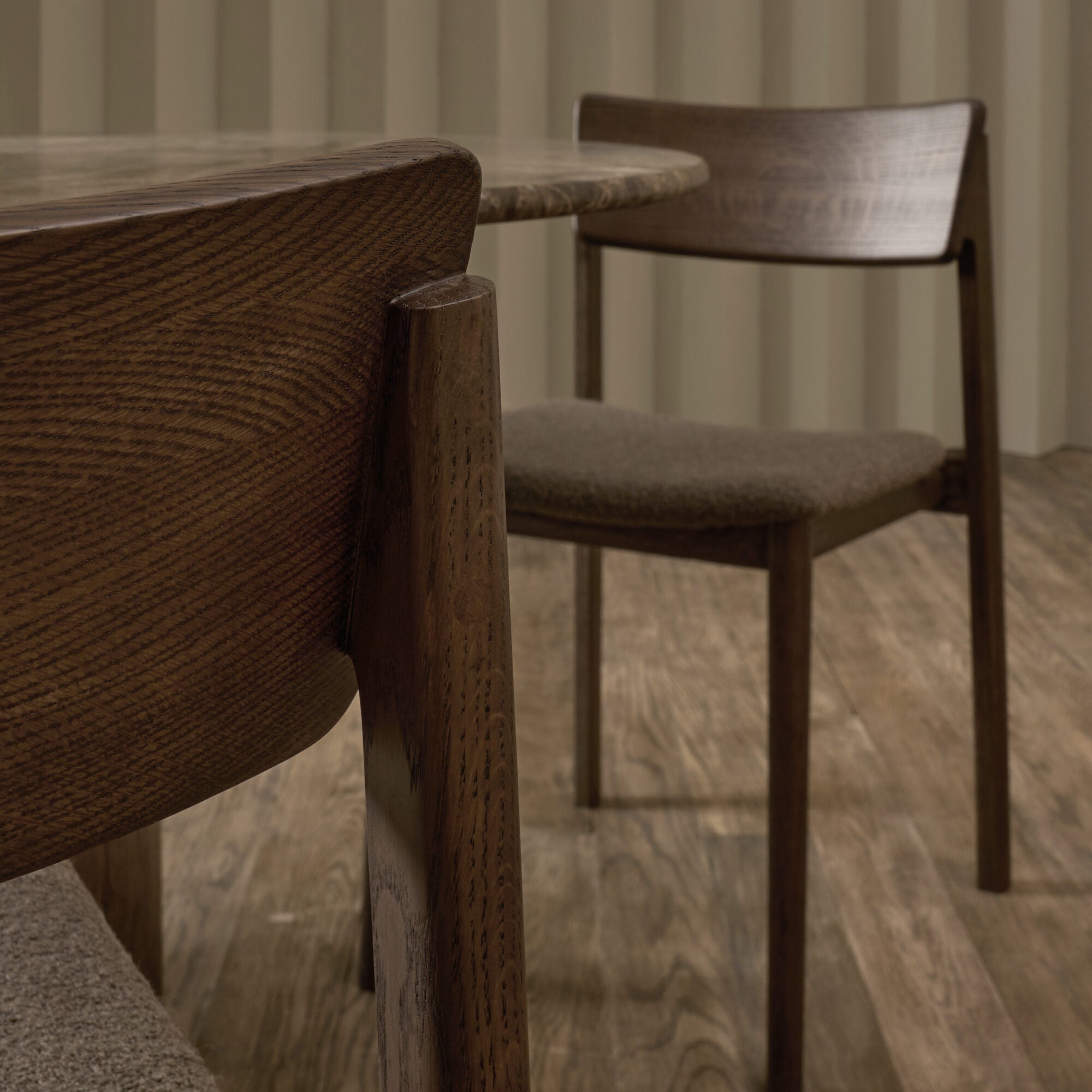 Close-up of two Wooden Stories Poise Dining Chairs in Espresso Oak with Fabric and a round table on a wood floor, set against neutral vertical paneling. The clean, modern lines and rich dark wood finish highlight contemporary style.