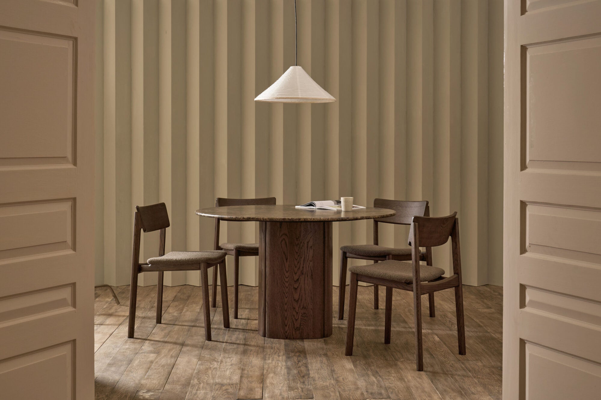 A round dark wood table with four Wooden Stories Poise Dining Chairs in Espresso Oak with Fabric sits on a wood floor. A white pendant lamp hangs above, with a striped beige wall behind, viewed through open double doors. Design by Patryk Koca.