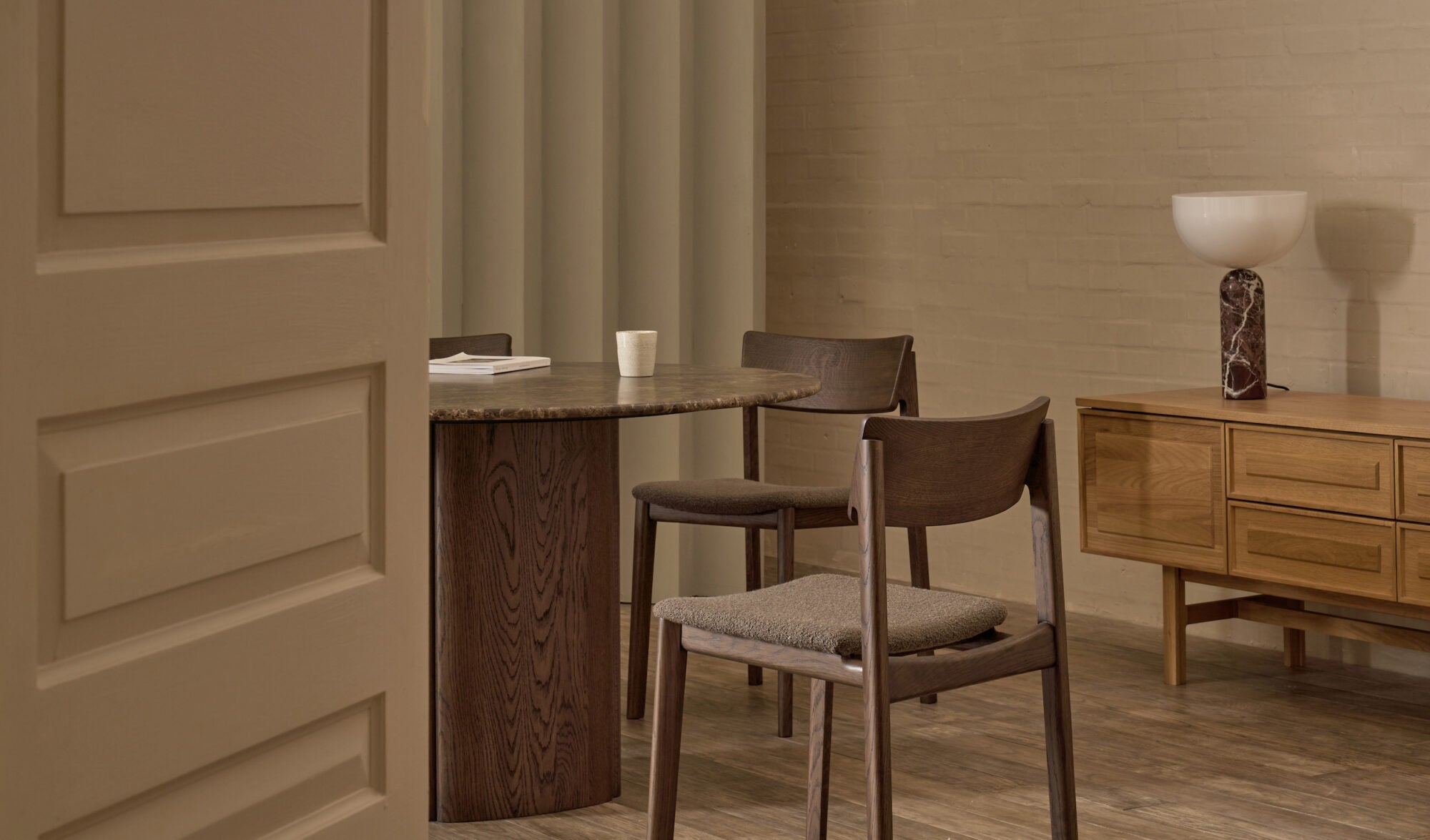 A minimalist dining room features a round dark wood table, three Wooden Stories Poise Dining Chairs in Espresso Oak with fabric, a sideboard, and a marble-based lamp. Neutral tones and soft lighting create a calm, modern atmosphere.