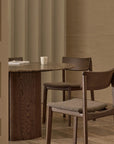 A minimalist dining room features a round dark wood table, three Wooden Stories Poise Dining Chairs in Espresso Oak with fabric, a sideboard, and a marble-based lamp. Neutral tones and soft lighting create a calm, modern atmosphere.