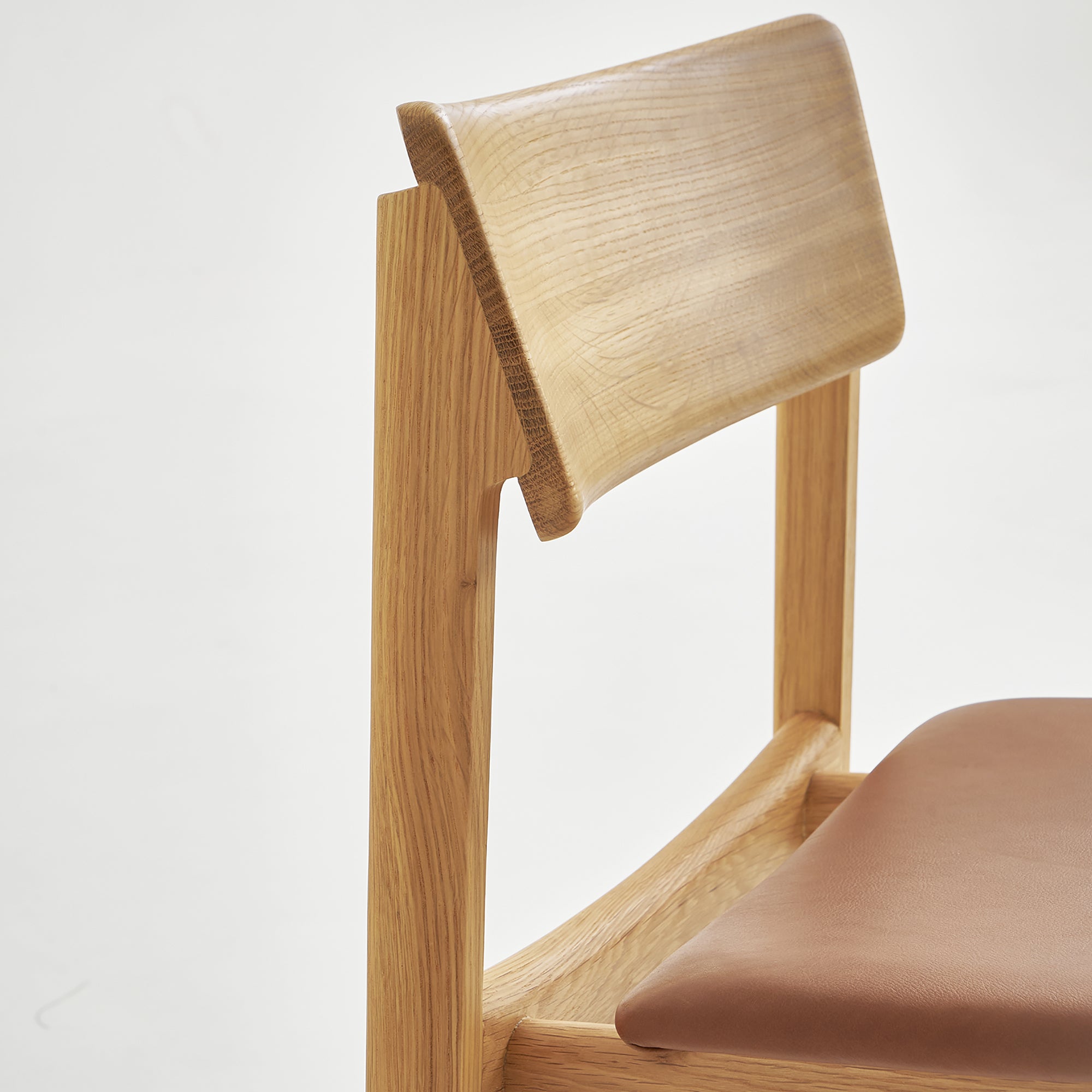 A close-up of Wooden Stories’ Poise Dining Chair in Light Oak with Leather highlights its modern Danish lines, curved backrest, and brown leather seat, set against a plain white background.