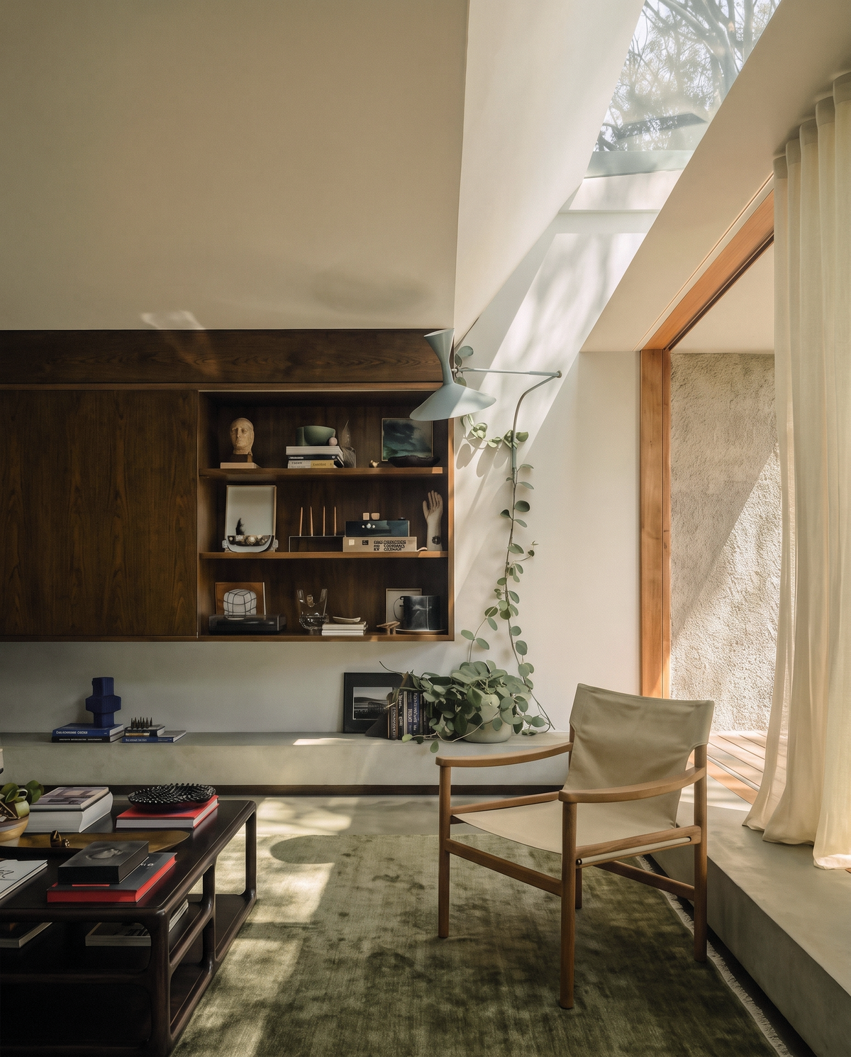 A cozy, sunlit living room featuring a green rug, wooden shelves filled with books and decor, the inviting Ethnicraft Safari Lounge Chair made of solid teak, abundant houseplants, and large windows adorned with sheer curtains that welcome in natural light.