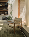 A sunlit modern living room features the Ethnicraft Safari Lounge Chair, a green rug, wall-mounted bookshelves, trailing plant, and sheer curtains. Warm natural light pours through a large window onto the scene.