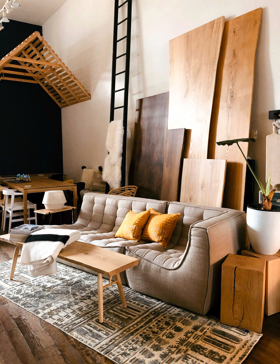 A cozy living room with a beige sofa, yellow pillows, wooden coffee table, patterned rug, and large wood panels leaning against the wall. A tall black ladder and dining area with chairs are in the background.