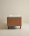 A light brown Sonder Sofa | Fabric with Leather Back by Wooden Stories, shown from the side with wooden legs, set against a plain beige wall and floor. This custom sofa features high-density foam cushions for superior comfort and lasting support.