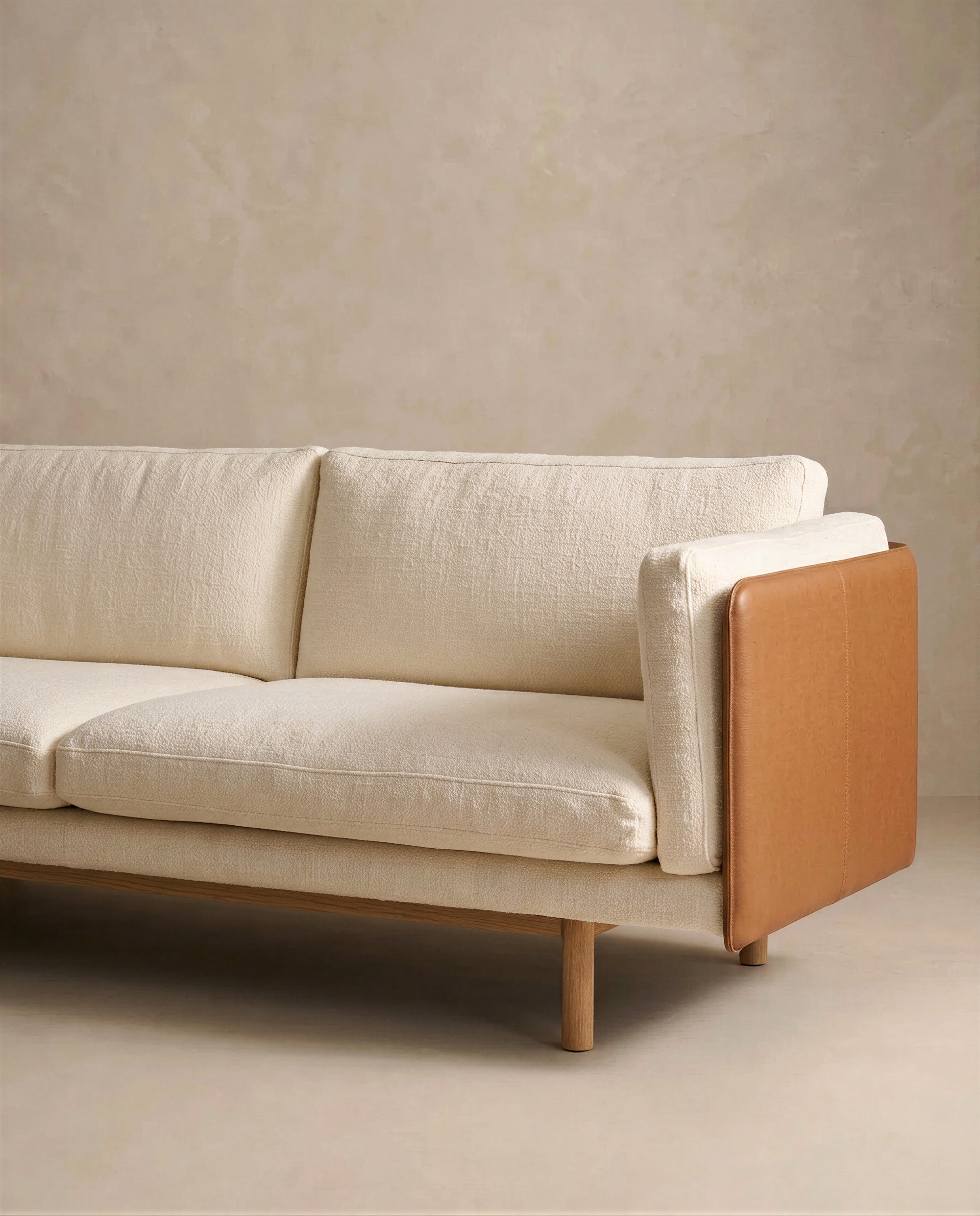 The Sonder Sofa | Fabric with Leather Back by Wooden Stories features modern cream-colored textured upholstery, wooden legs, and a tan leather panel on the armrest from the Danish collection, set against a neutral beige wall and floor.