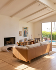 A bright living room featuring the customizable Sonder Sofa | Fabric with Leather Back by Wooden Stories, a cozy fireplace, captivating artwork, wooden desk, and expansive windows overlooking a pool and lush greenery. Neutral tones and abundant natural light evoke a modern yet inviting atmosphere inspired by the Danish collection.
