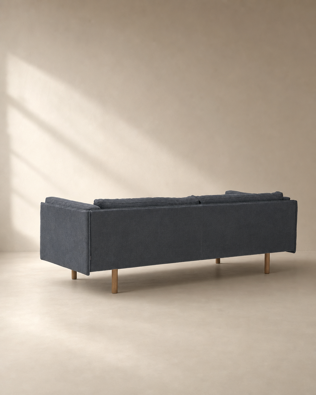 A Sonder Sofa | Fabric by Wooden Stories in blue-gray, featuring wooden legs from the Danish collection, is shown from the back in a minimalist beige room where sunlight streams through a window and casts shadows on the wall.