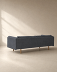 A Sonder Sofa | Fabric by Wooden Stories in blue-gray, featuring wooden legs from the Danish collection, is shown from the back in a minimalist beige room where sunlight streams through a window and casts shadows on the wall.