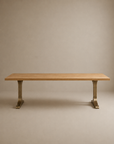 The Tommaso Dining Table by SOBU features a rectangular, modern design with a smooth solid wood surface and two brass T-shaped metal pedestal legs, showcased against a plain beige background.