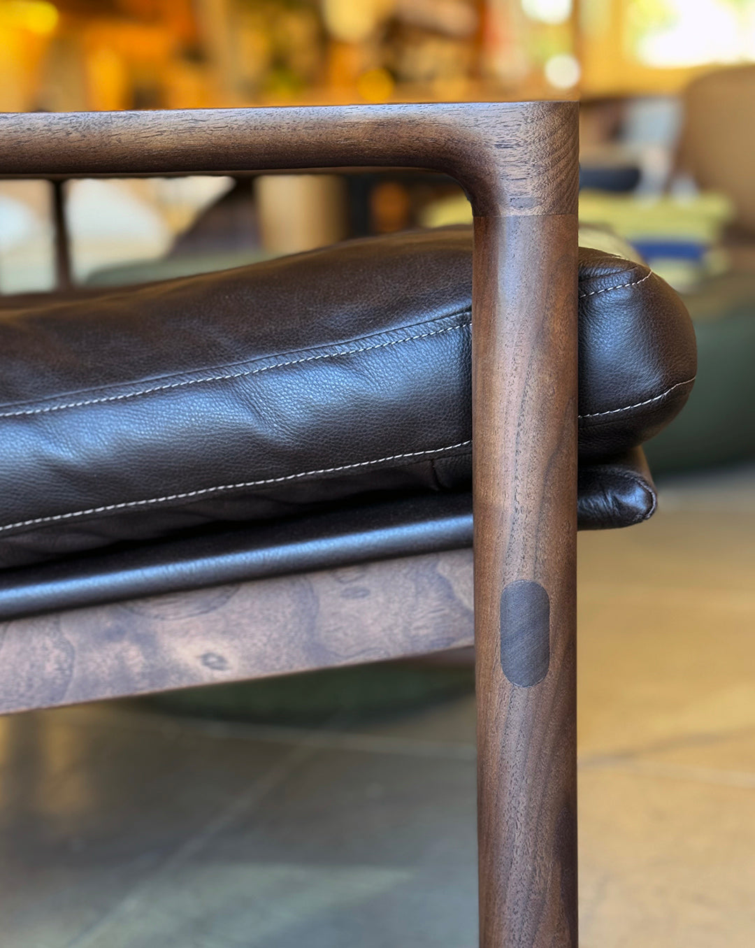 Close-up of the Troubadour Lounge Chair | Leather by Wooden Stories, featuring a dark leather cushion on a smooth, rounded wood frame with rich grain, set in a softly blurred, warmly lit indoor scene.