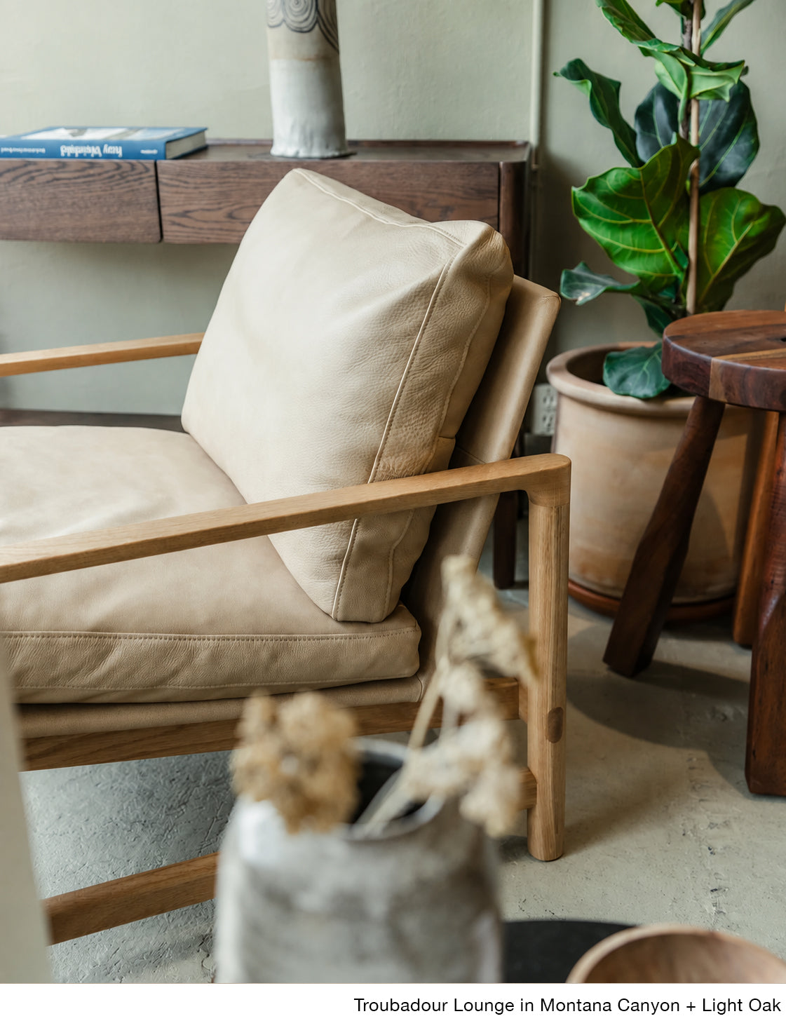 The Troubadour Lounge Chair | Leather by Wooden Stories in light tan leather and walnut sits beside a potted plant, wooden stool, and console table to create a cozy, modern living space.