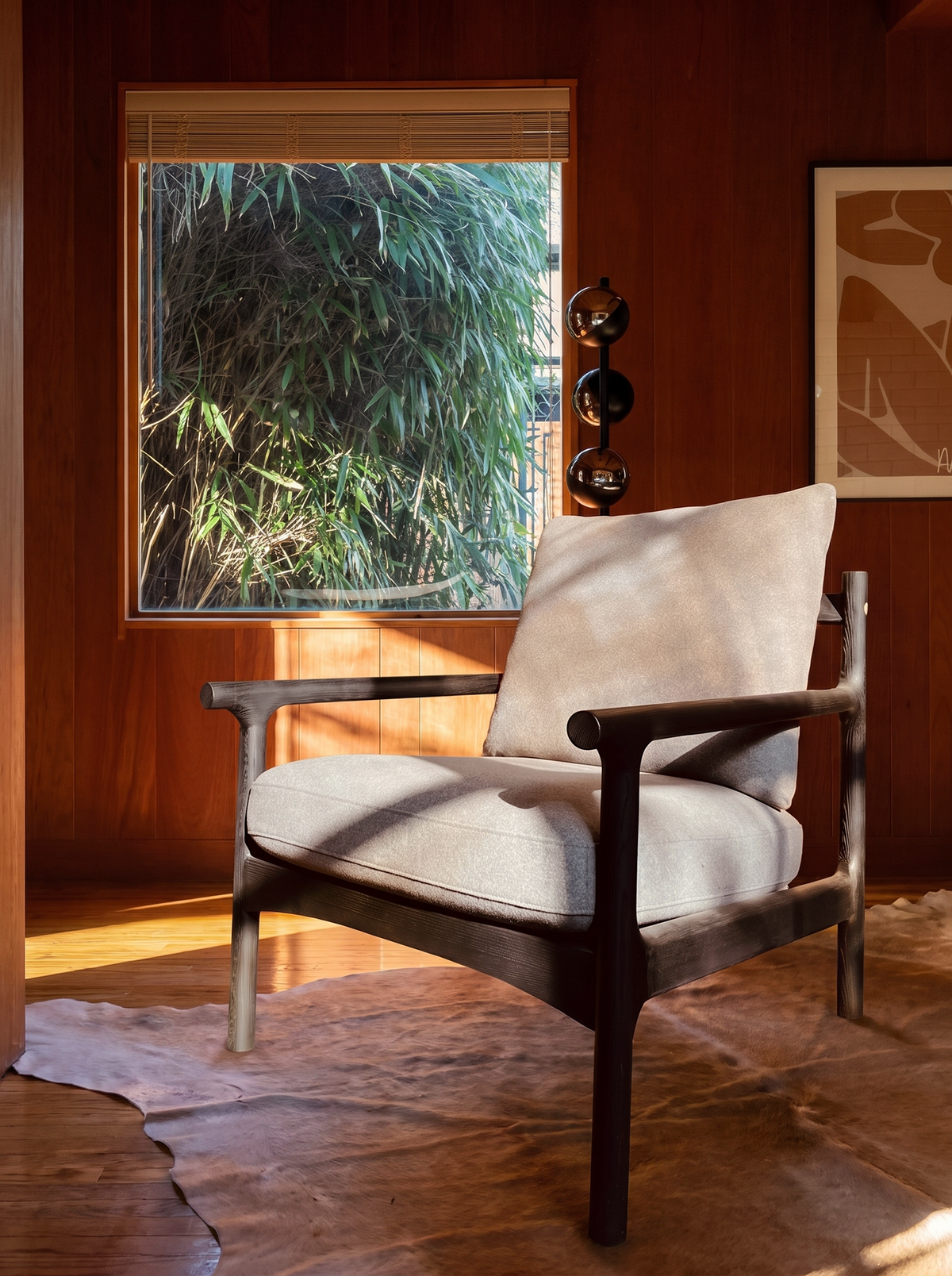 A SOBU Viking Lounge Chair - Black Ash sits on a cowhide rug in a sunlit room with wood-paneled walls, a large window framed by bamboo, and abstract wall art.
