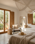 Sunlit bedroom with large windows, neutral decor, a canopy bed, abstract wall art, cozy armchair, wooden accents, the Mars Terra Side Table by SOBU as a minimal nightstand; greenery outside and a straw hat on the bed.