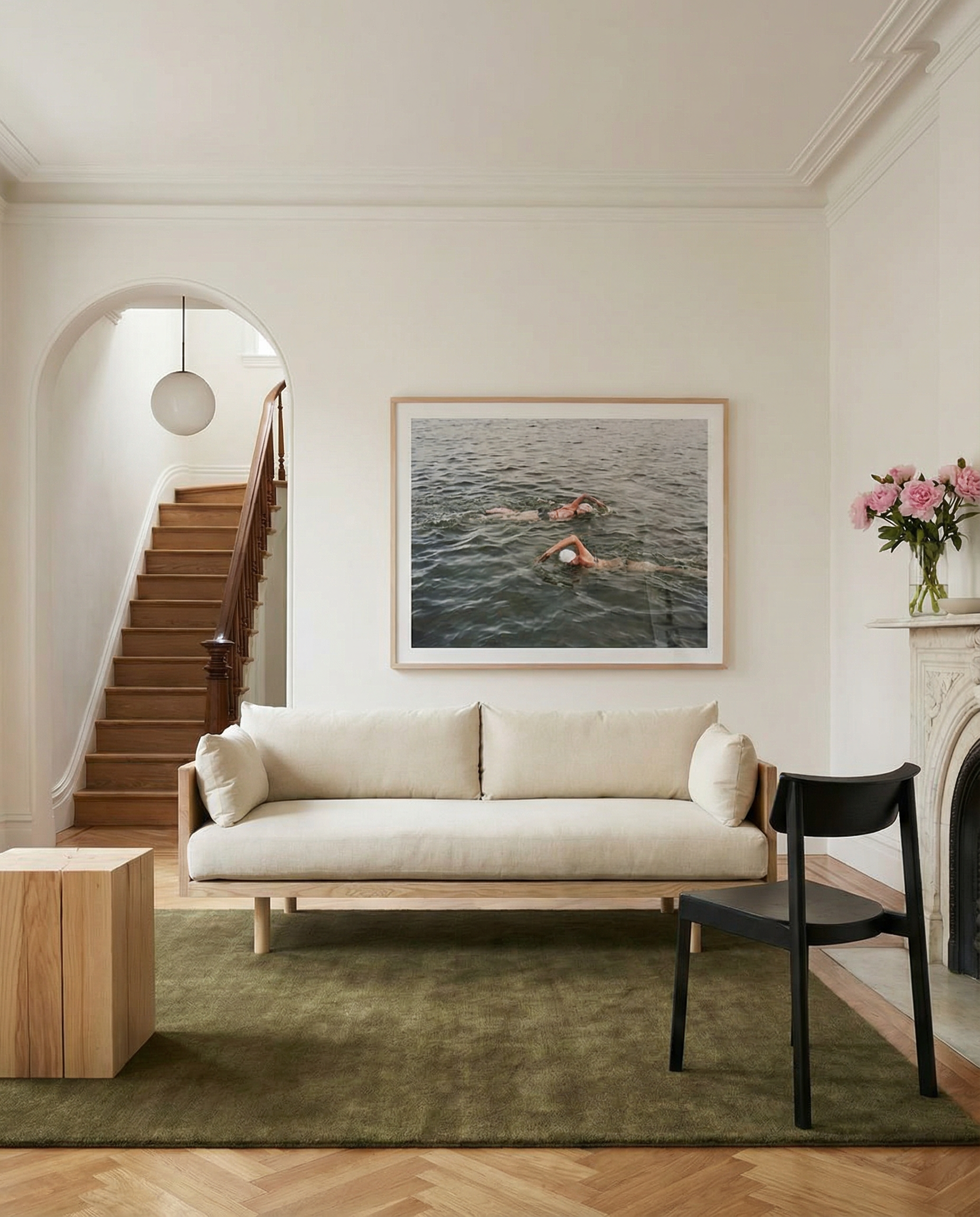 A modern living room showcases the SOBU Woody Sofa in white ash with natural kapok cushions, a black chair, wooden side tables, and a green rug. Above the sofa hangs a swimmers photo. A staircase, arched doorway, and fireplace with pink flowers complete the scene.