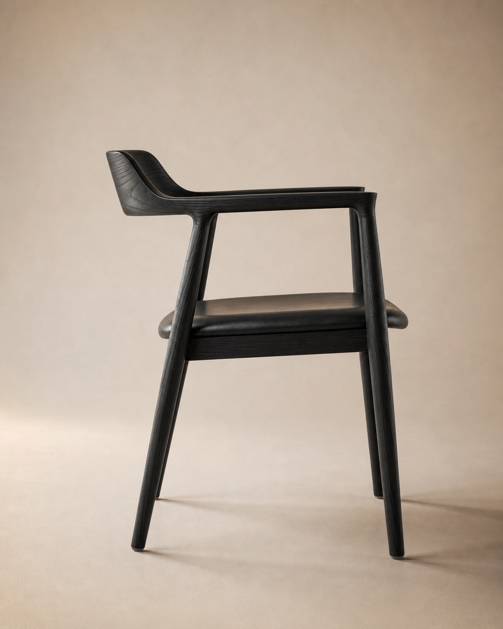 The SOBU Sven Dining Chair in black ash features armrests, a curved backrest, and sleek, modern lines. Shown in side profile against a beige background, it’s optionally upholstered in black Italian leather for a refined finish.