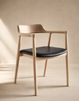 The SOBU Sven Dining Chair in White Ash and leather features a curved backrest, armrests, and a black cushioned seat, crafted with a natural finish and shown against a neutral beige background with soft lighting.