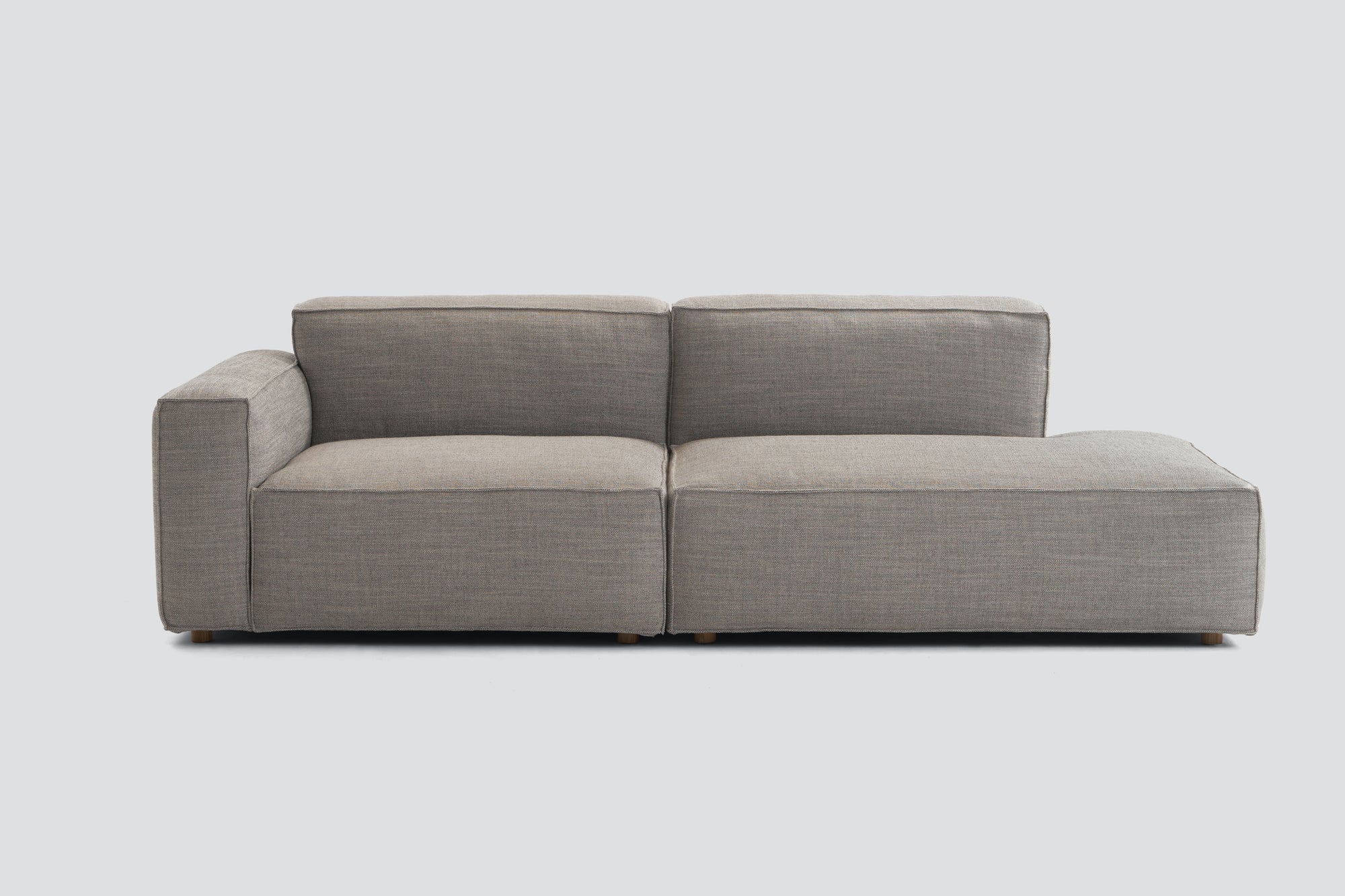 The Baker Modular Sofa | Fabric • M1 by Wooden Stories is a modern, light gray sofa with a left armrest, two seat cushions, and a right chaise lounge, set against a plain, light background.