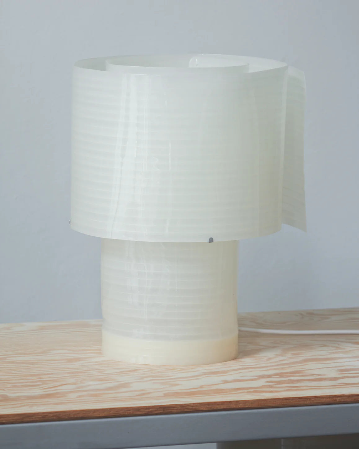 The Snegl Scallop Table Lamp by RUSSO BETAK STUDIO features a sculptural, 3D-printed cylindrical design with an overlapping translucent shade, offering a soft glow and minimalist elegance inspired by the sea.