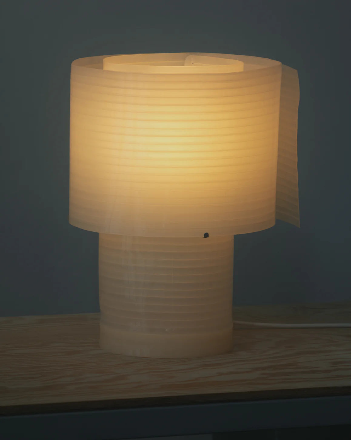 The Snegl Scallop Table Lamp by RUSSO BETAK STUDIO sits on a light wooden surface, its ribbed, biodegradable design casting a soft, elegant glow—bringing a story from the sea against a dark gray backdrop.